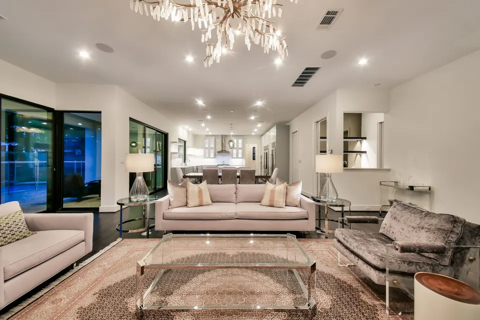 Open-plan living room with cream sofas, dark wood flooring, a chandelier, black-framed glass doors, and a kitchen beyond